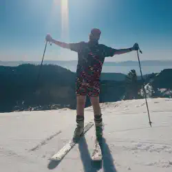 Short shorts and matching pattern button up shirt with a bright colored hat standing in front of Lake Tahoe