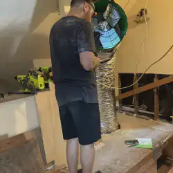 Alex wiring the fan in the attic. The fan can be seen in the background attached to lumber. The duct is already attached to the fan.